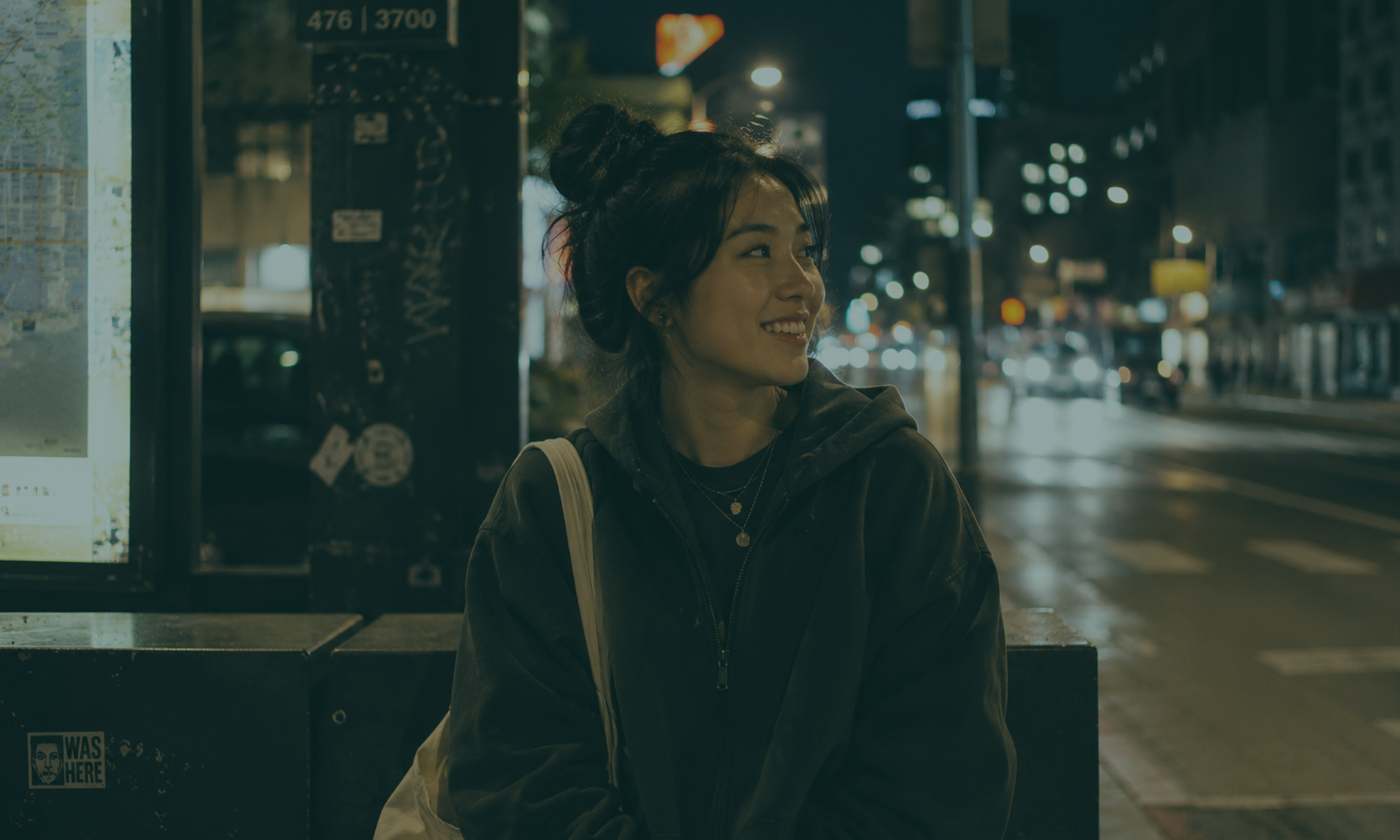 Young woman smiling on a city street at night surrounded by glowing lights and an urban backdrop.
