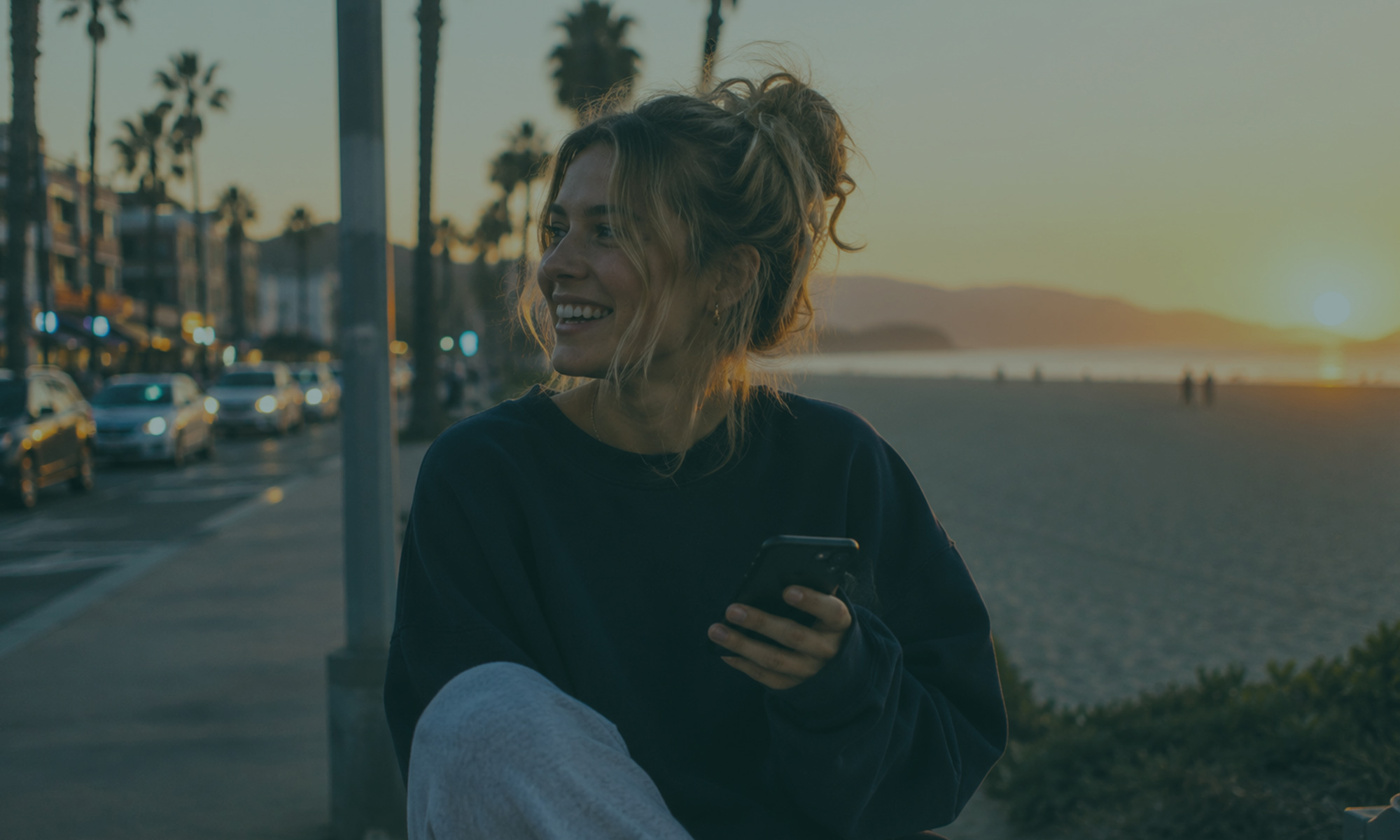 Young woman smiling with her phone near the beach at sunset, enjoying a relaxed moment by the ocean.